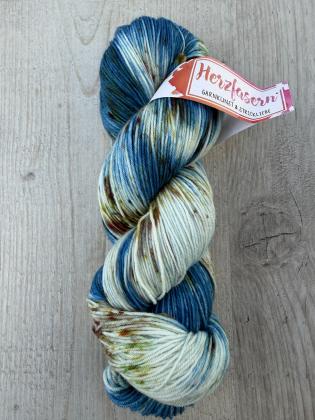 Handgefärbter Garnstrang in kühlen Blau- und Petroltönen mit cremefarbenen Abschnitten und feinen Sprenkeln in warmen Farben auf hellem Holzhintergrund.